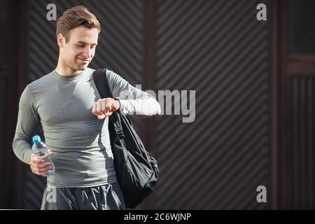 Der junge lächelnde Mann geht mit Sporttasche zum Training und in Sportkleidung, schaut auf Tracker zur Hand Stockfoto