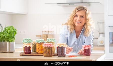 Gut aussehende Frau mit einem Stift und Notizbuch von hausgemachten Eingemachtes in der Küche umgeben. Stockfoto