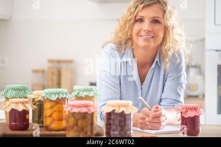 Gut aussehende Frau mit einem Stift und Notizbuch von hausgemachten Eingemachtes in der Küche umgeben. Stockfoto