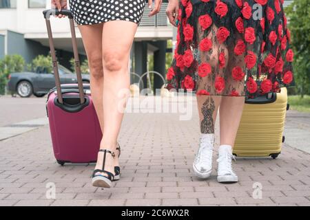 Von unten zugeschnittenes Foto von zwei eleganten Touristenfrauen, die mit Koffern auf der blumigen Straße der Stadt spazieren. Stockfoto