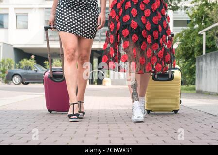 Von unten zugeschnittenes Foto von zwei eleganten Touristenfrauen, die mit Koffern auf der blumigen Straße der Stadt spazieren. Stockfoto