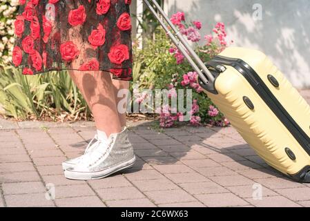 Von unten zugeschnittenes Foto einer Touristenfrau in Blumenkleid und Tenis-Schuhen, die mit ihrem Koffer auf der blumigen Straße der Stadt spazieren. Stockfoto