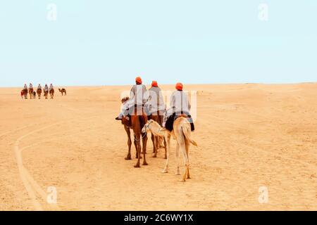 Touristen in gestreiften Gewändern und orangen Turbanen auf ihren Köpfen, die auf zweiköpfigen Kamelen für einen Spaziergang in der Sahara, Tunesien, reiten Stockfoto