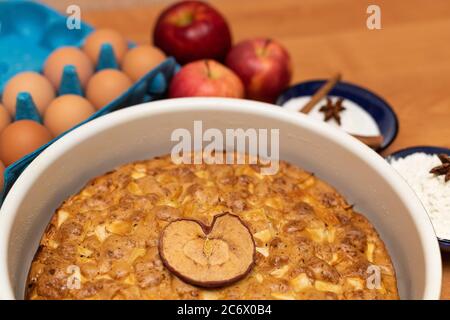 Apfelkuchen mit herzförmigem Apfel auf Holztisch mit Backzutaten Stockfoto