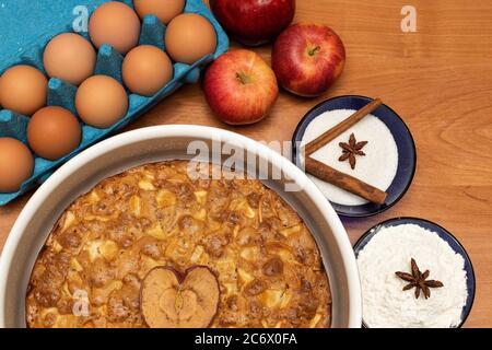 Apfelkuchen auf einem Holztisch mit Backzutaten: Äpfel, Eier, Zucker, Mehl, Zimt und Badian Stockfoto