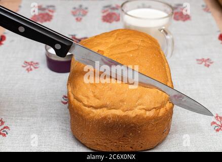 Messer schneidet Weizenbrot, eine Tasse Milch auf einem Leinentischtuch Stockfoto