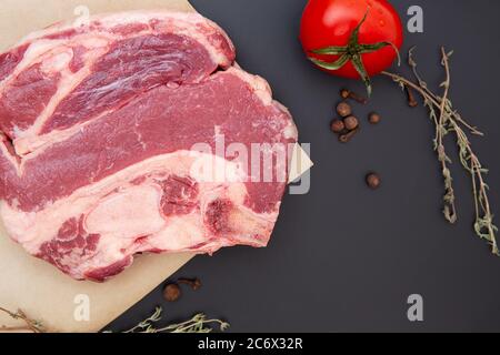 Frisches rohes Rindersteak mit Gewürzen, Kräutern und Gemüse. Das Fleisch ist auf dem Knochen. Thymian, schwarzer Pfeffer, Tomaten, Rindfleisch auf schwarzem Stein. Hintergrund für das Essen. Stockfoto