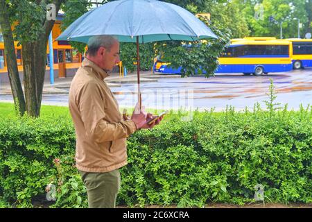 Ein Mann, der am Busbahnhof steht und einen Regenschirm auf einen Bus wartet. Mann wartet auf einen Bus, SMS auf Handy an einem regnerischen Tag. Regnerischer Tag in einem Stockfoto