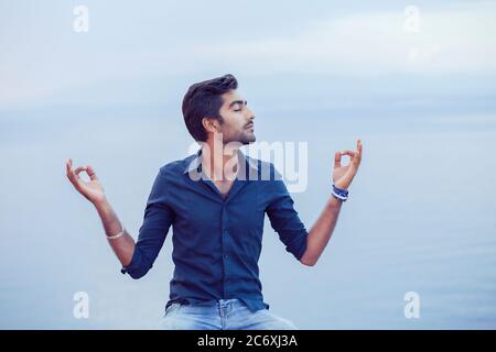 Mann in Yoga-Meditation Pose sitzt auf einem Felsstein Ufer über dem Meer unter tiefen Atem entspannen bei Sonnenuntergang blauen Himmel und Meer auf Hintergrund. Suchen Sie har Stockfoto