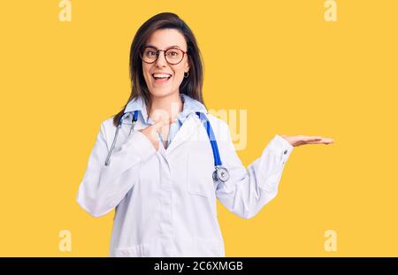 Junge schöne Frau trägt Arzt Stethoskop und Brille erstaunt und lächelt an der Kamera, während mit der Hand und zeigt mit dem Finger. Stockfoto