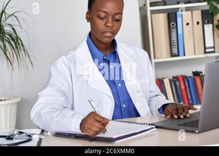 afroamerikanischer Arzt, der Krankenakte in Notebook mit Laptop am Schreibtisch schreibt. Stockfoto