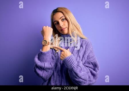 Junge schöne blonde Frau trägt lässige Rollkragenpullover über lila Hintergrund in Eile zeigt, um Zeit zu sehen, Ungeduld, Blick auf die kam Stockfoto