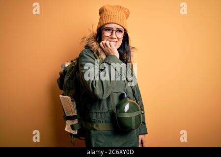 Junge Wanderfrau trägt Wanderrucksack, Kantine und Karte auf gelbem Hintergrund gestresst und nervös mit Händen auf Mund beißende Nägel. Anxie Stockfoto