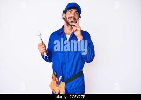 Schöner junger Mann mit lockigen Haaren und tragen Baumeister Jumpsuit Uniform und halten Schraubenschlüssel ernstes Gesicht denken über Frage mit Hand auf Kinn Stockfoto