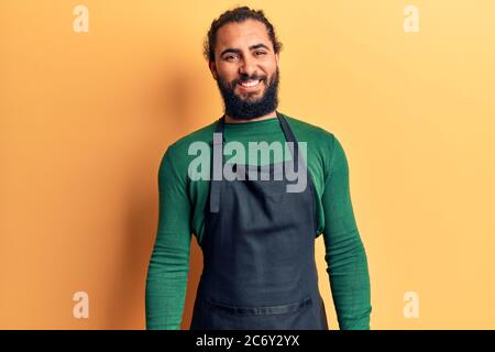 Junger arabischer Mann mit Friseurschürze, der positiv und glücklich stehend und lächelnd mit einem selbstbewussten Lächeln und Zähnen aussieht Stockfoto