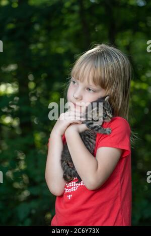 Ein fünfjähriges Mädchen, das im Sommer ein tabby Kätzchen im Freien mit Bäumen im Hintergrund hält. Stockfoto