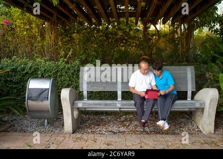 Ältere chinesische Paar auf einer Bank sitzen und lesen ein mobiles Tablet zusammen Stockfoto