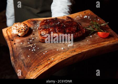 Auf einem Holzbrett liegt ein appetitliches, frisch zubereitetes gegrilltes Fleischsteak. Mit Salzstücken bestreut, daneben gibt es einen Rasmarin und eine Tomatenscheibe. Stockfoto