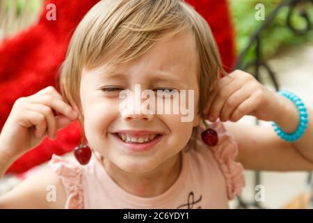 Ein kleines Mädchen lächelt und hält Kirschen an ihre Ohren, als wären sie Ohrringe Stockfoto