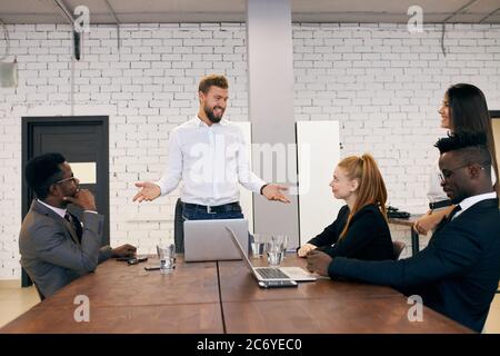 Ausländische Leute halten Konferenz im Business Center in modernen Büro. Internationales Team hört kaukasischen bärtigen Mann in weißem formelles Hemd, usi Stockfoto