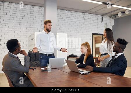 Ausländische Leute halten Konferenz im Business Center in modernen Büro. Internationales Team hört kaukasischen bärtigen Mann in weißem formelles Hemd, usi Stockfoto