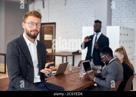 Schöner junger Mann in Smoking und Brillen mit Smartphone am Arbeitsplatz, während seine Kollegen im Hintergrund sprechen Stockfoto