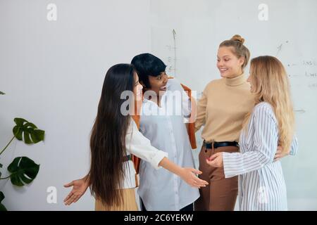 Junge schöne kaukasische und afrikanische Damen tragen lässige Kleidung umarmen einander feiern ihren Sieg in Projekt. Weiß moderner Bürohintergrund Stockfoto