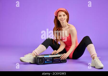 Junge, fröhliche, sportliche Frau mit roten Haaren und flexiblem, sportlichen Körper sitzt nach dem Fitnesstraining auf dem Boden und schaltet ihren tragbaren Audioplayer aus. Stockfoto