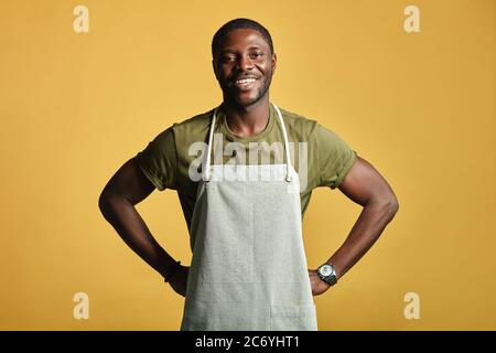 Afrikanischer Bauer mit muskulösen Händen und sportlichem Körper, in Schürze gekleidet, lächelnd auf Kamera mit toothy Lächeln auf gelbem Hintergrund. Die Menschen Bekennen Sich Stockfoto