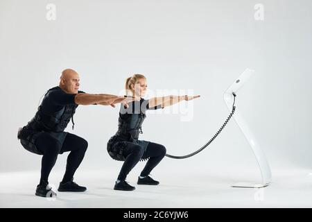 Ein Paar trägt Anzüge für Elektromuskelstimulation EMS Durchführung Kniebeugen. Volle Länge Seitenansicht Foto. Copy space.family ist gern Fitness Stockfoto