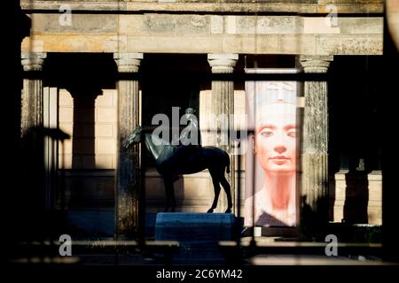 Berlin, Deutschland. Juli 2020. Im Neuen Museum ist ein Plakat der Nofretete und die Bronzeskulptur 'Amazonas zu Pferd' des deutschen Bildhauers Louis Tuaillon im Licht der Morgensonne zu sehen. Nach zweijähriger Evaluierung der Stiftung Preußischer Kulturbesitz (SPK) wird der Wissenschaftsrat heute auf einer Pressekonferenz seine Empfehlung zur Auflösung der Dachorganisation staatlicher Museen, Bibliotheken und Institute erläutern. (Fotografiert durch eine Glasscheibe) Quelle: Christoph Soeder/dpa/Alamy Live News Stockfoto