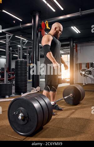 Haarlose brutale männliche Athlet auf den Füßen in der Nähe von Barkeeper in der professionellen Turnhalle, Vorbereitung auf Deadlift als Teil des Fitness-Programm, bodybuil zu machen Stockfoto