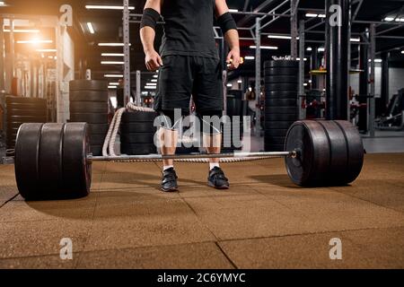 Nahaufnahme der Hände des Powerlifters, die in der Nähe einer schweren Langhantel in schwarzen Sneakers stehen, weiße Socken tragen, Zeit zum Training in der Sporthalle, Innenaufnahme Stockfoto