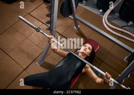 Schöne sportliche Frau, die Hand auf Eisenstange auf der roten Bank unter schwerer Langhantel liegend hält, versucht, Gewicht zu heben, mit konzentriertem Gesicht aufsehend, ta Stockfoto