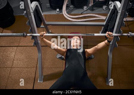 Schöner sportlicher Gewichtheber auf Bank in schwarzer stylischer Sportbekleidung liegend, Hände hochheben, um Eisenstange zu halten, Training im modernen Fitness-Center mit h Stockfoto