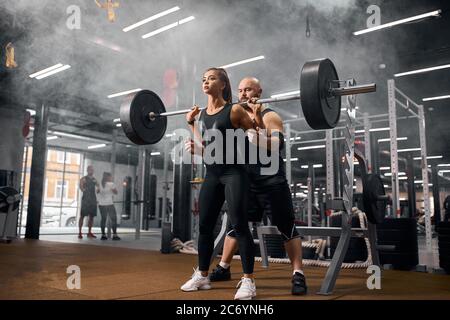 Junge sportliche Frau konzentriert sich auf das Heben schwere Langhantel, dabei Kniebeugen mit ruhigen starken Gesicht, Ausdruck von Vertrauen in die Turnhalle, Training zusammen mit haarlos Stockfoto