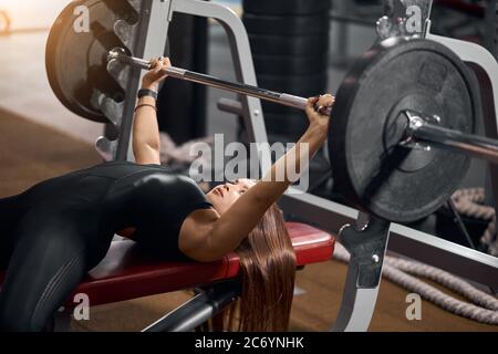 Sportliche junge Kraftheber mit langen geraden Haaren, unter eiserner Langhantel liegend, sich im Rahmen des Fitnessprogramms für das Gewichtheben vorbereiten, Seite s. Stockfoto