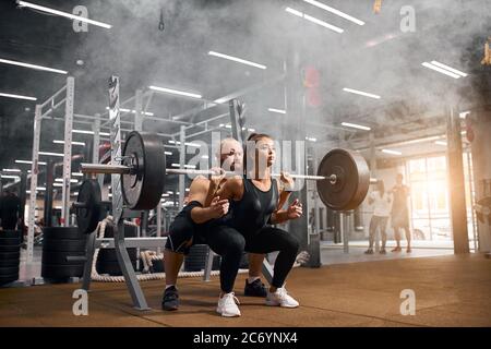 Unhöfliche brutale Trainer mit kahlem Kopf und dicken Brünette Bart hinter jungen weiblichen Powerlifter stehen, sie unterstützen, Kontrolle Prozess der Hocke Stockfoto