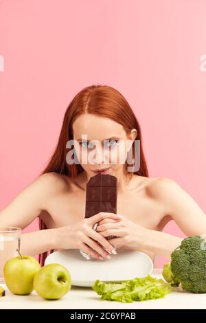 Glücklich überglücklich magere Frau mit ungesunder Haut und Taschen unter ihren Augen, brechen ihre Ernährung, essen mit Freude eine Tafel von dunkler Schokolade, sitzen shir Stockfoto