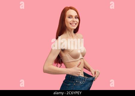 Slim Happy girl in großen Jeans isoliert auf rosa, zeigt ihre schlanke Figur nach Diät und Training isoliert auf rosa Hintergrund Stockfoto