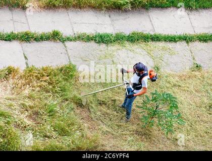 Ein Servicemitarbeiter mit einem benzinbetriebenen Trimmer mäht hohes Gras auf dem Rasen eines Stadtparks entlang eines Fußgängerwegs. Platz kopieren, Draufsicht. Stockfoto