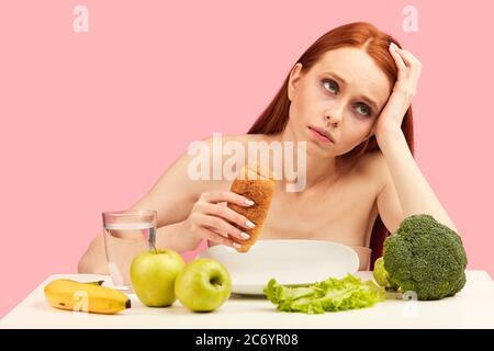 Gelangweilt gleichgültig apathischen rothaarigen Frauen in BH essen Croissant starrte in den Raum ignorieren rohe Früchte über rosa Hintergrund. Stockfoto