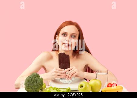 Glücklich überglücklich magere Frau mit ungesunder Haut und Taschen unter ihren Augen, brechen ihre Ernährung, essen mit Freude eine Tafel von dunkler Schokolade, sitzen shir Stockfoto