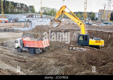 Raupenbagger in Prozess der Verladung Sand Boden auf Multi-Tonnen-Bergbau-LKW auf Baustelle mit Kräne Gebäude und Arbeiter im Hintergrund. Stockfoto