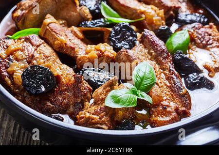 Schweinebrippen-Tajine-Eintopf mit Pflaumen, Rotwein, Brühe, Zwiebel und Knoblauch, mit frischem Basilikum verziert, serviert auf einer schwarzen Backform auf rustikalem Holzrücken Stockfoto