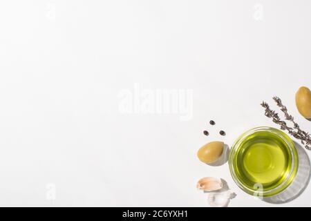 Ansicht von oben Olivenöl in der Schüssel neben grünen Oliven, Knoblauch, Kräuter und schwarzem Pfeffer auf weißem Hintergrund Stockfoto