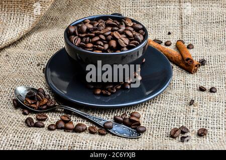 Schwarzer Kaffeebecher voll von organischem Kaffee Bohnen und Zimtstangen auf Leinen - Bild Stockfoto