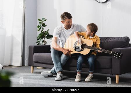 Vater und Sohn schauen sich gegenseitig an, während Junge lernen, wie man akustische Gitarre spielt Stockfoto