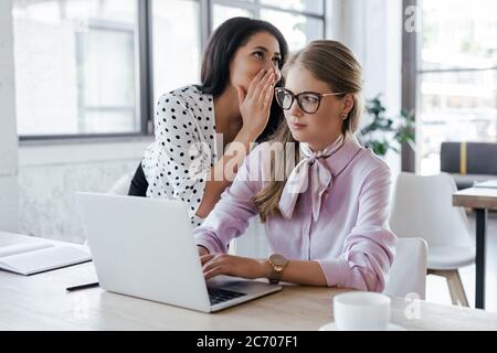 Geschäftsfrau flüstert in Brille vor dem Ohr des Kollegen, während sie im Büro klatscht Stockfoto