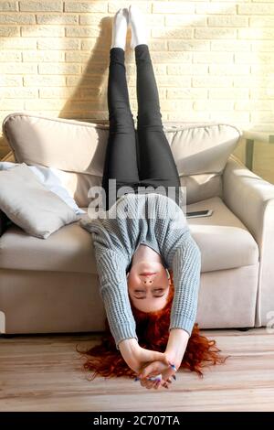 Fröhliches rothaariges Teenager-Mädchen hört Musik auf dem Sofa auf dem Kopf. Zu Hause Sonne auf der Mauer. Stockfoto